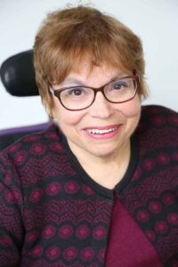 A headshot of Judy Heumann, a cis-gender white woman who is a wheelchair user with short brown hair. She is wearing red glasses and a maroon and black embroidered sweater with the top buttoned with a matching maroon shirt underneath. She is smiling kindly.