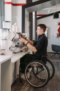 A barista in a wheelchair, representing disabled entrepreneurs, prepares coffee at a modern café counter.