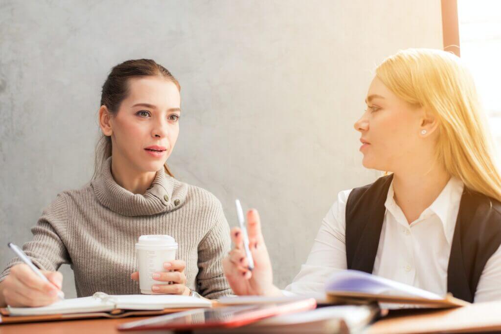 Two women sit at a table, chatting and jotting notes about a career-focused moving guide; one holds a coffee cup while the other gestures with a pen, planning for a smooth transition.