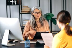 Smiling woman with gray hair interviews another woman at an office desk with documents and a computer.