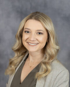 Smiling woman with wavy blonde hair in a beige blazer poses in front of a gray background.
