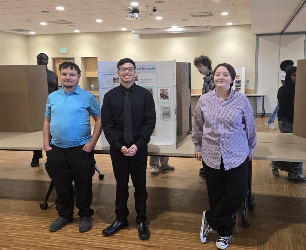 Three people stand in front of a project display board at an indoor event with others in the background.