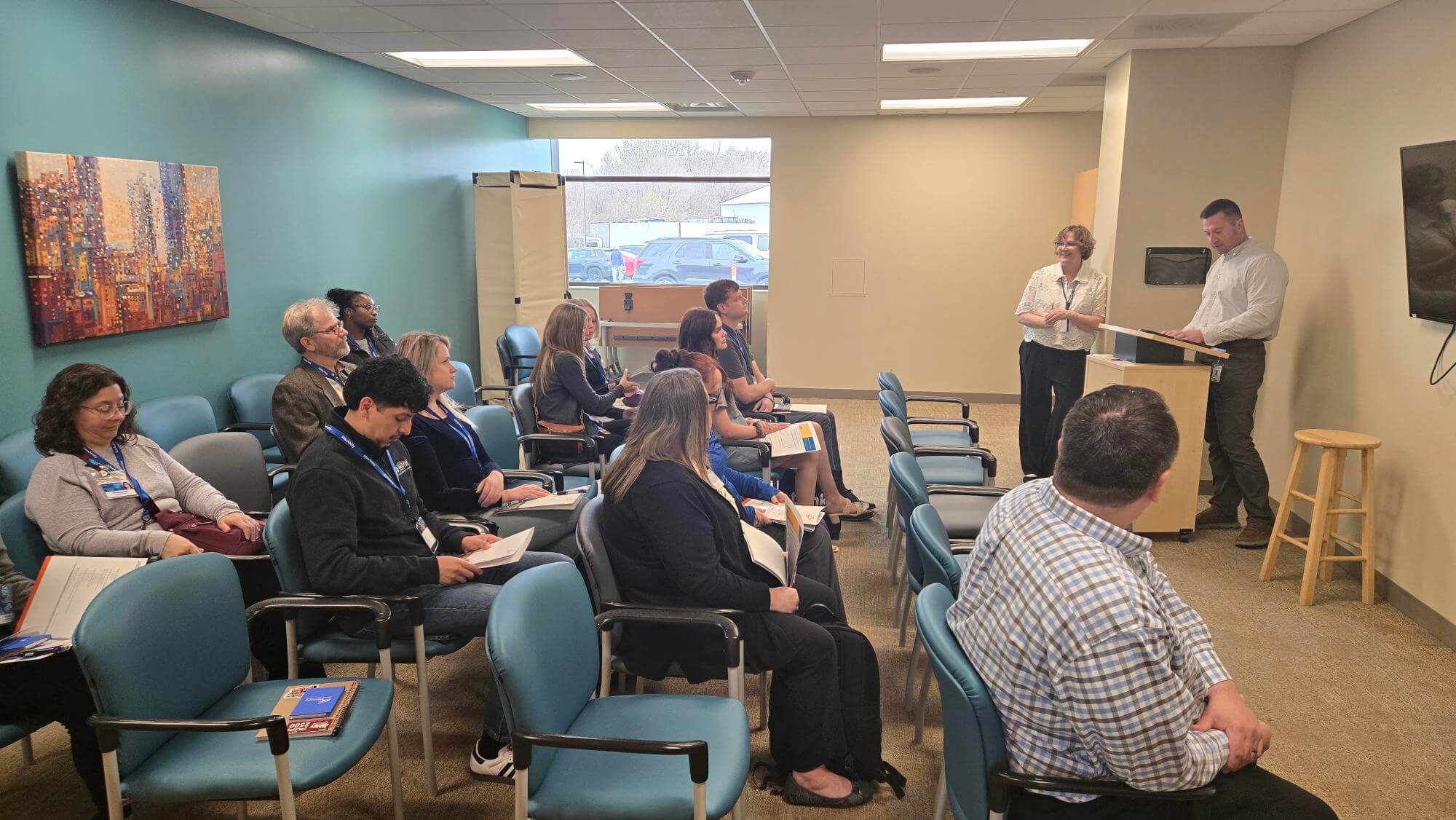 People seated in a small conference room listen to two presenters at the front near a podium and screen.