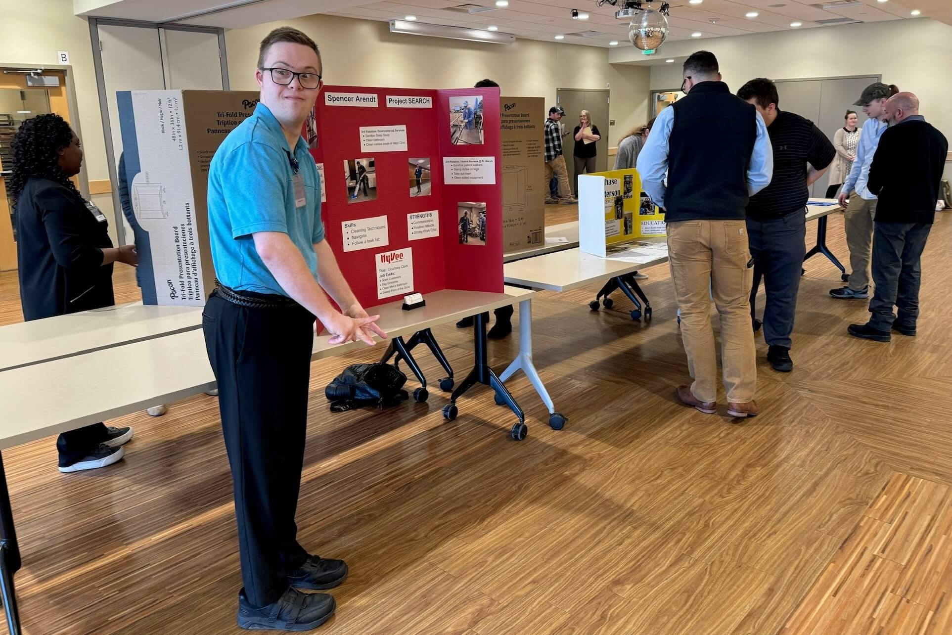 A man in a blue shirt presents a science project display board at a science fair with several people attending.