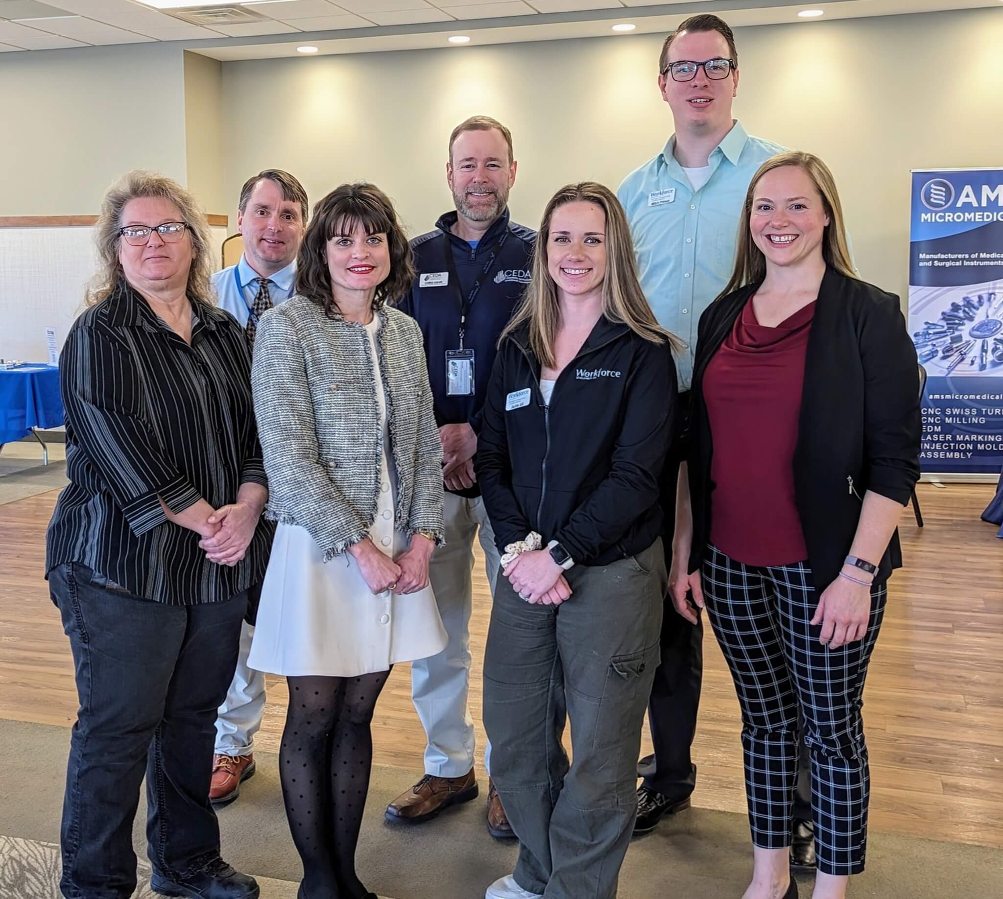 Seven people pose and smile together indoors at a professional event with banners in the background.