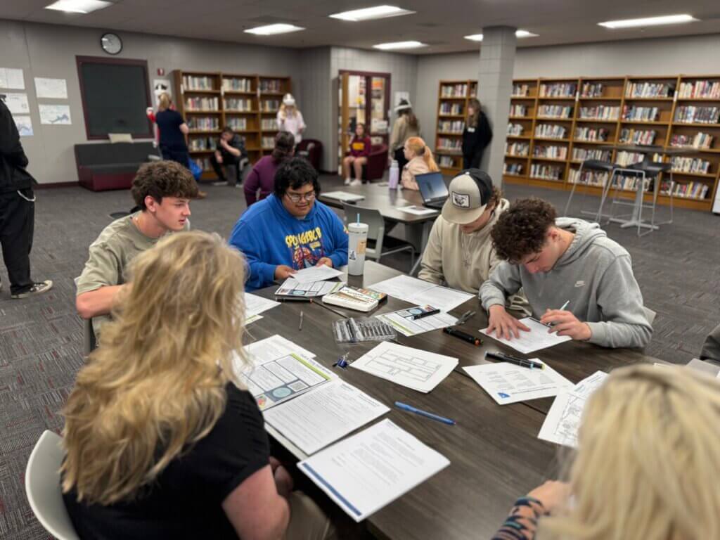 Young adults sit at a table in a library, working together on worksheets and projects.