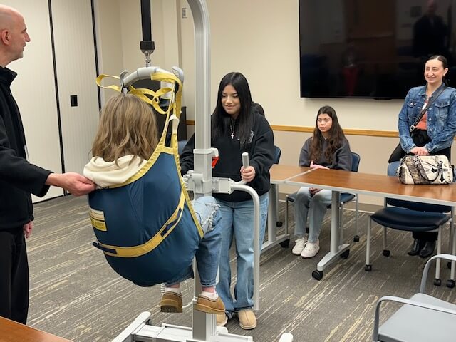A girl operates a patient lift, raising another girl, while three young adults observe in a classroom.