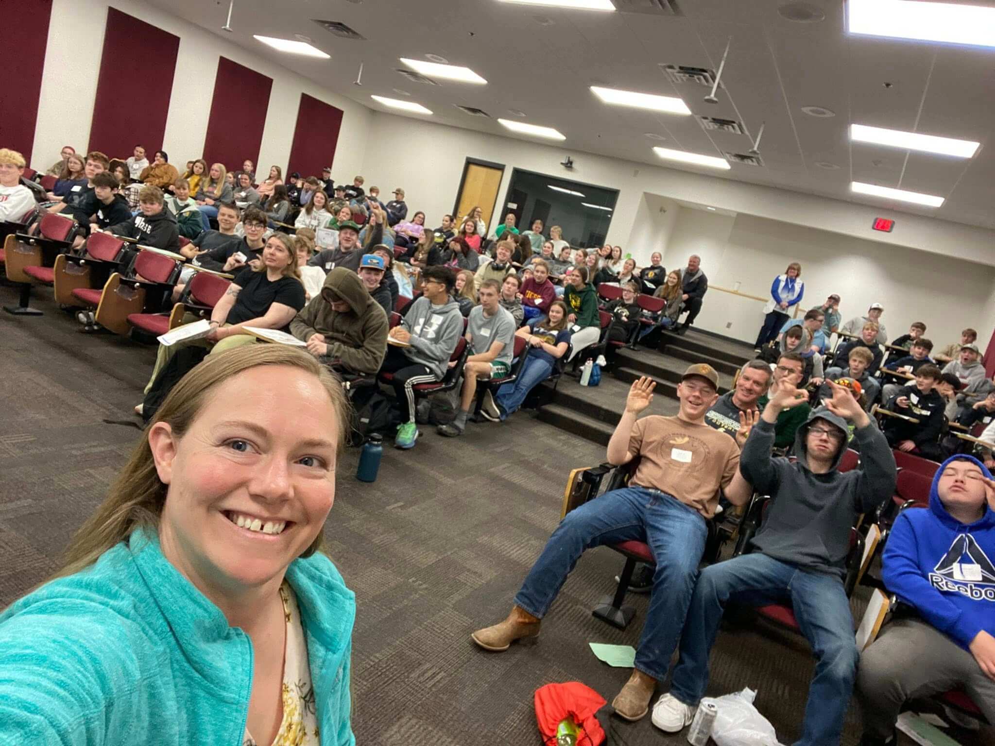 A woman takes a selfie with a large group of young adults seated in an auditorium, many youth smiling and waving enthusiastically.