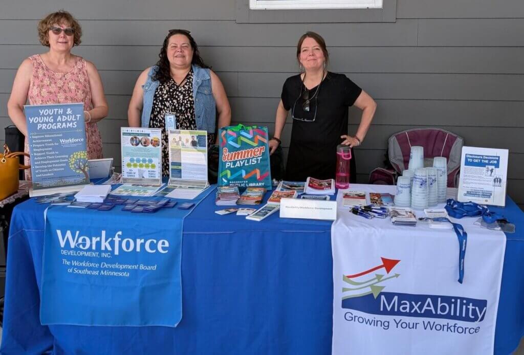 Three women stand behind a table with workforce and youth program materials at an outdoor event.