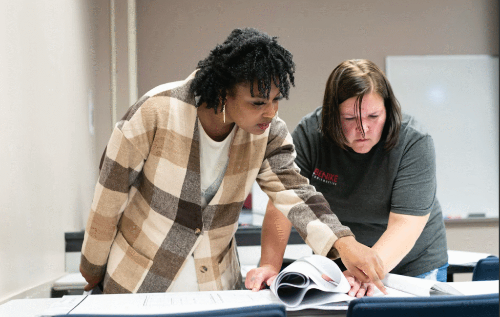 Two women stand and look at papers on a table, collaborating in a classroom or office setting as part of the Bridges to Careers program.