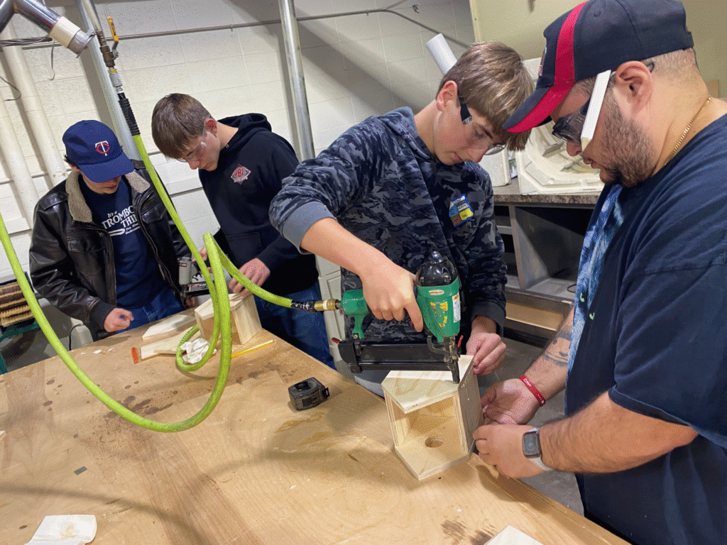 Four young adults assemble wooden boxes with tools at a workbench in a workshop.