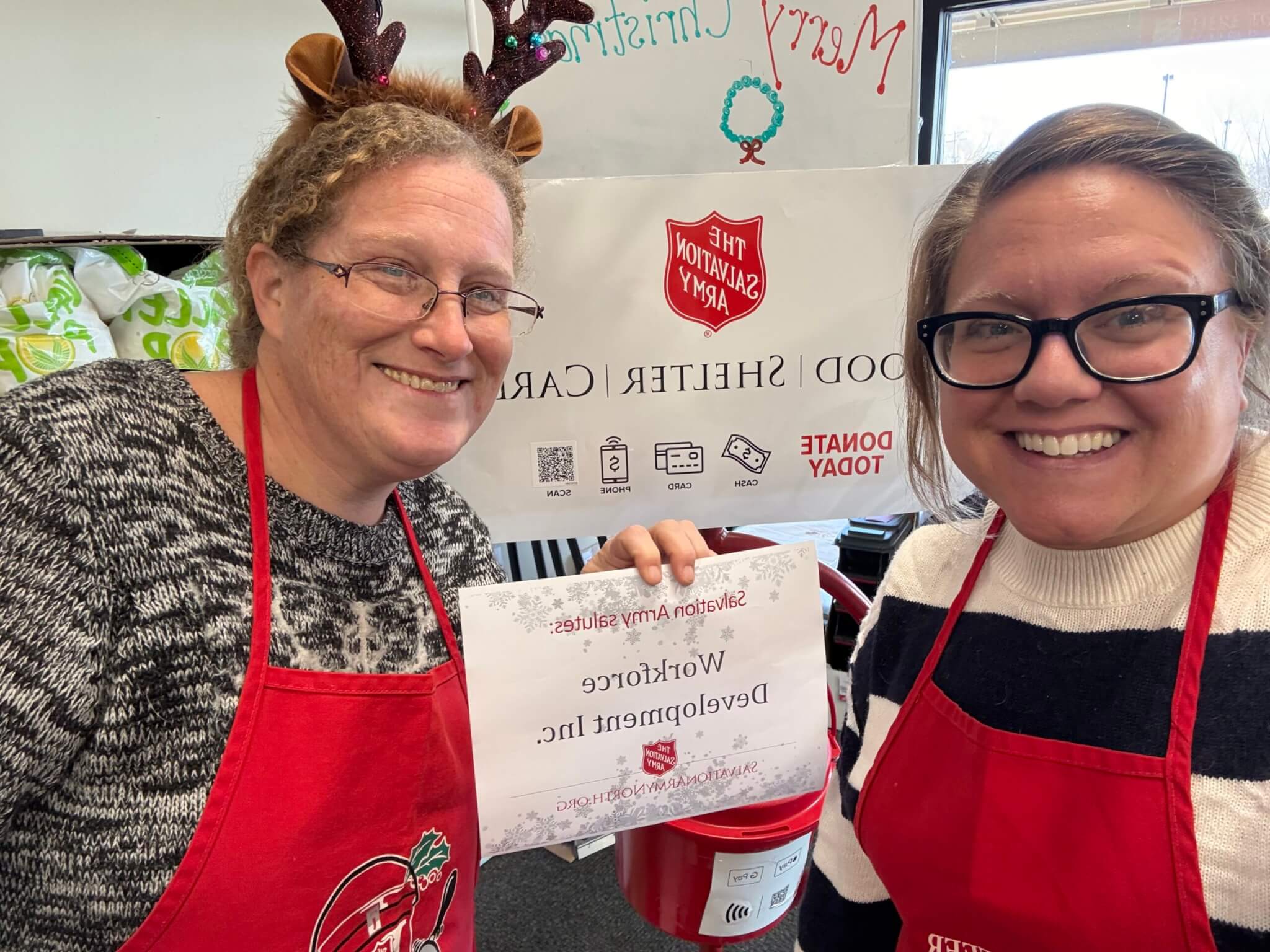 Two smiling women in aprons at a Salvation Army donation site, holding a Workforce Development sign.