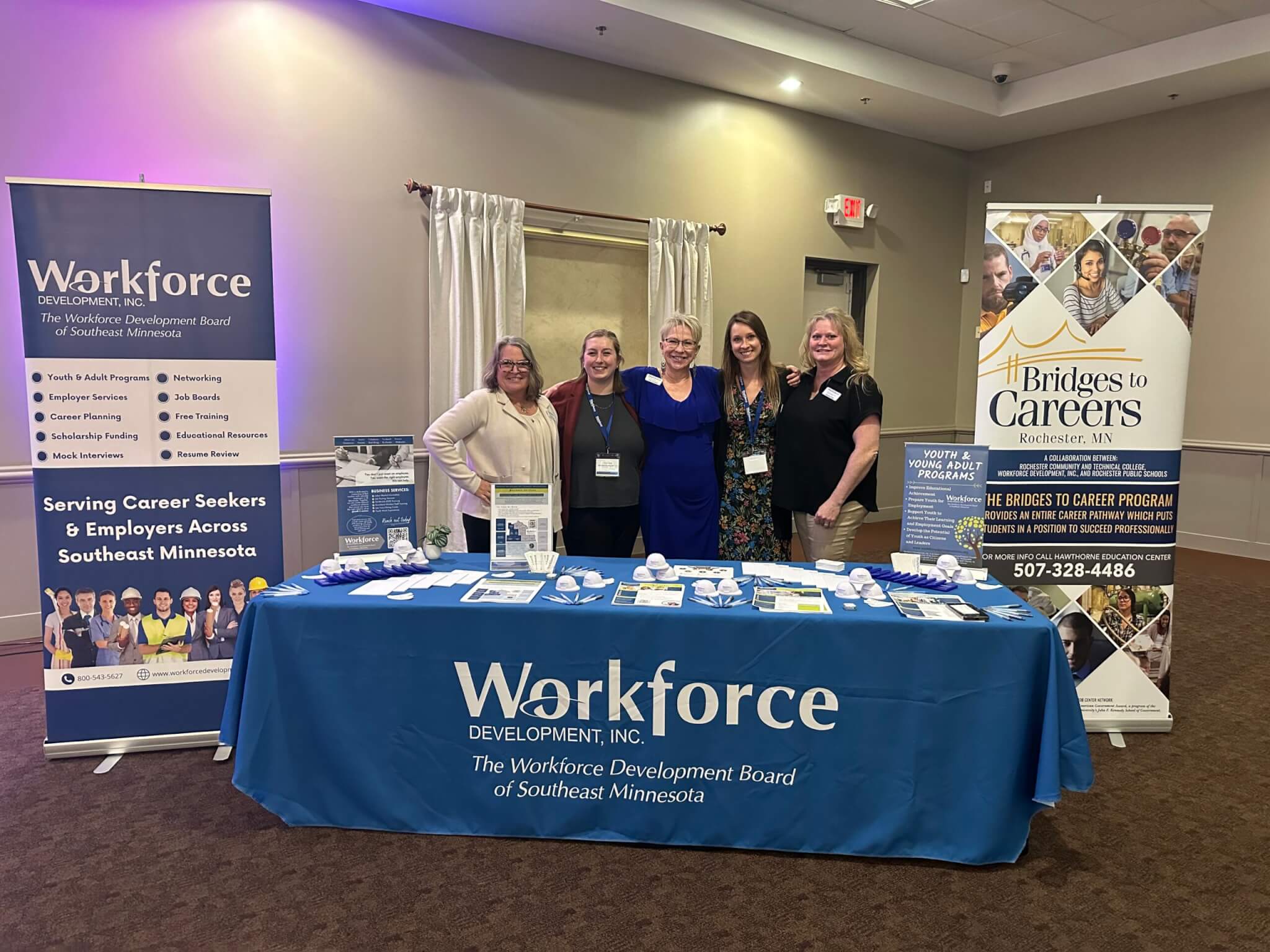 Five women stand behind a Workforce Development info table at an indoor event, smiling at the camera.