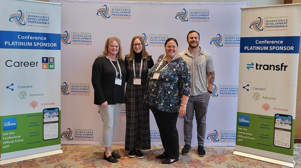 Four people stand smiling in front of a Workforce Development Professionals conference banner, celebrating their shared commitment to workforce development.