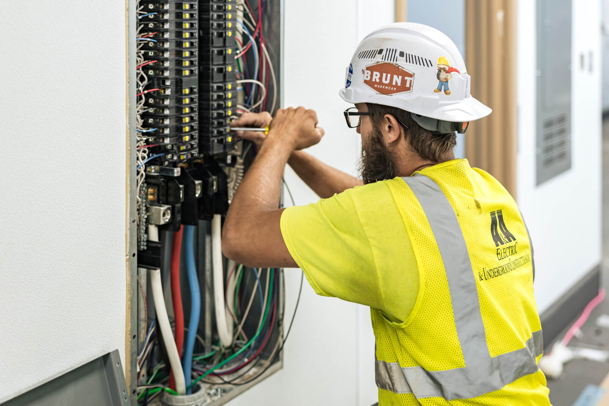 Electrician in safety gear working on a circuit breaker panel with exposed wires in a building.