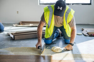 A worker in a yellow vest spreads adhesive on a floor with a trowel while installing flooring.
