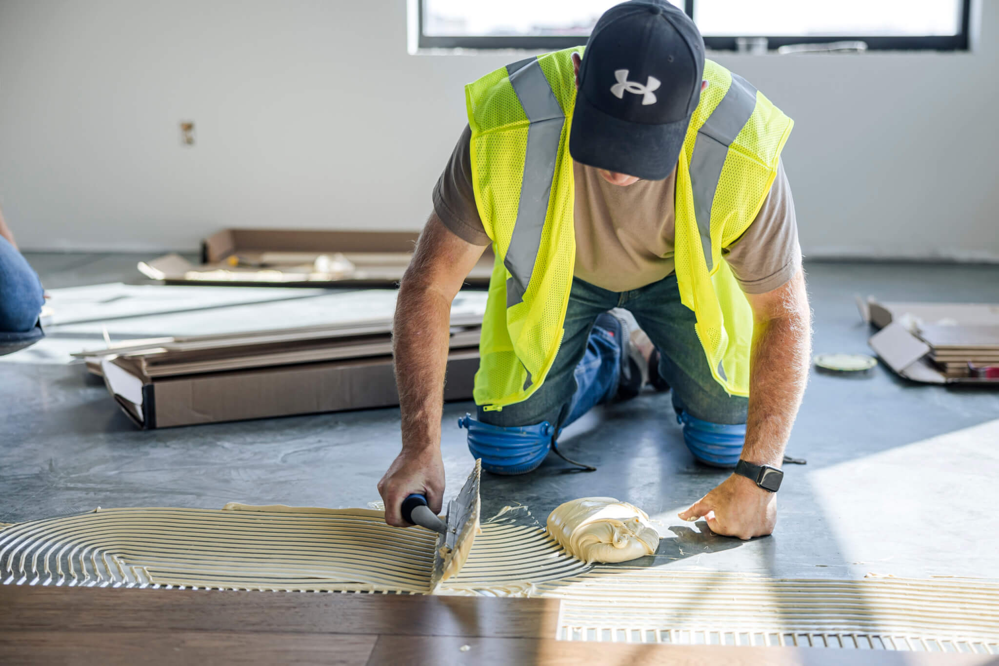 A worker in a yellow vest spreads adhesive on a floor with a trowel while installing flooring.