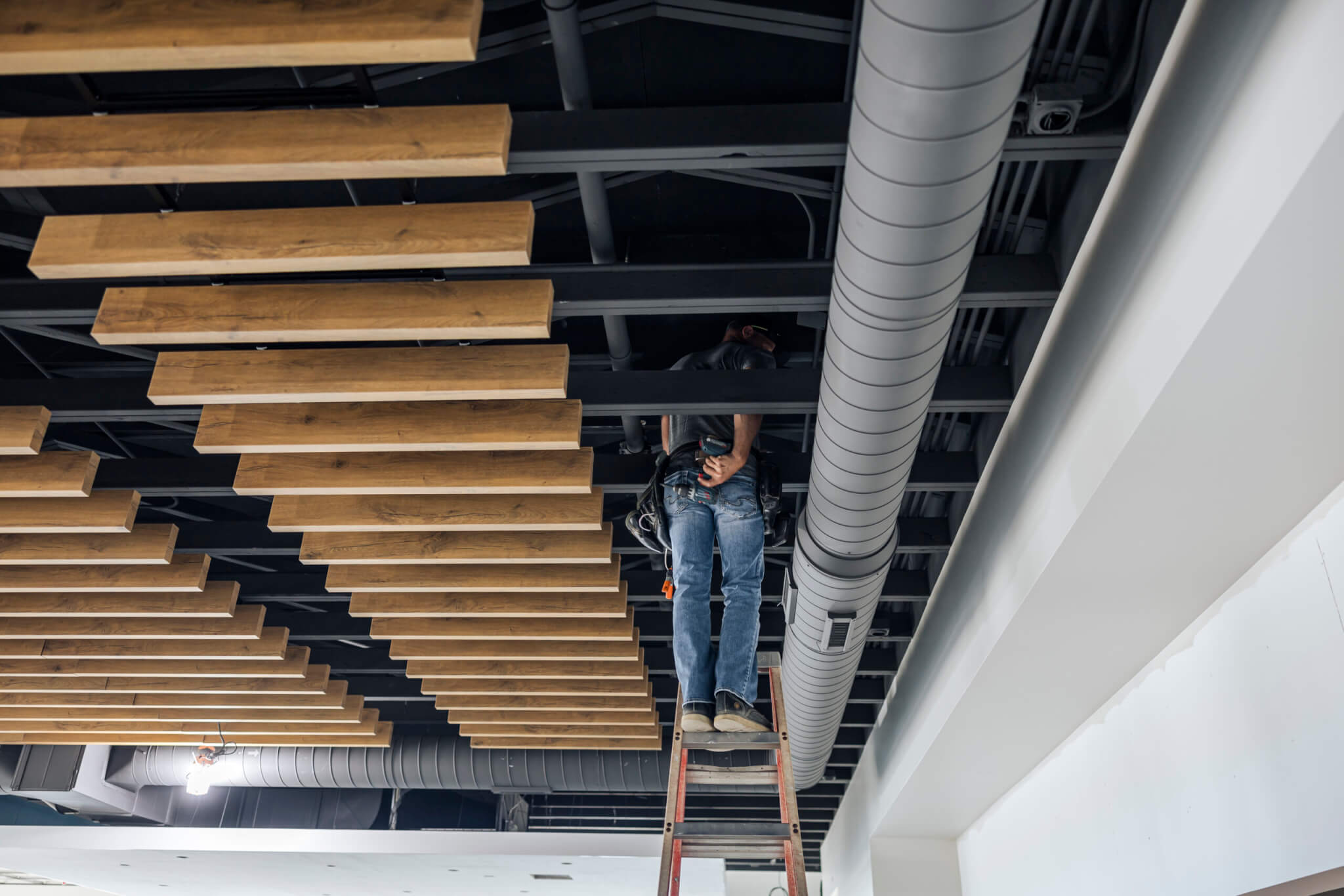 A worker stands on a ladder fixing a ceiling with exposed pipes and wooden beams.