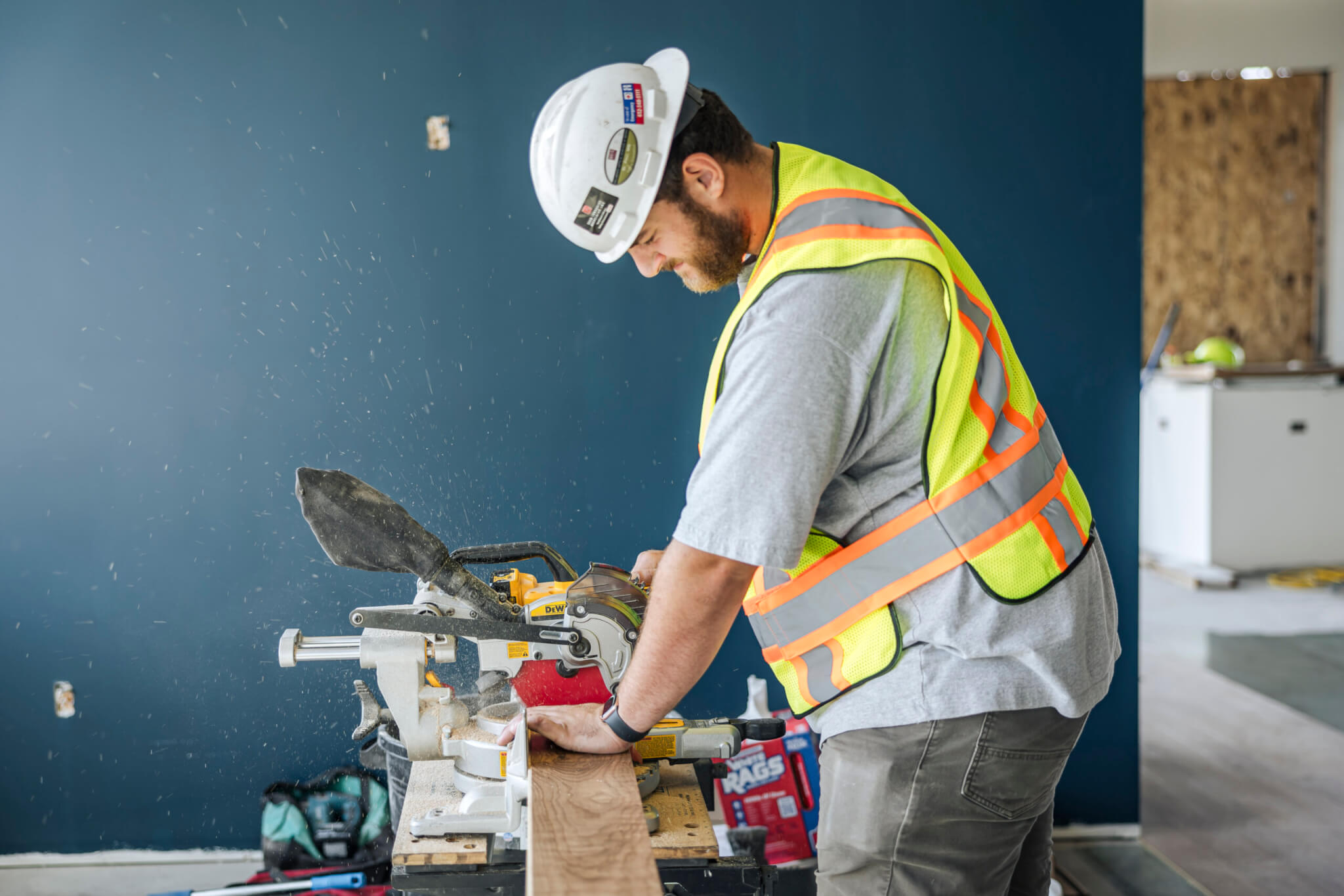 Construction worker in safety gear cutting wood with a power saw indoors; blue wall in background.