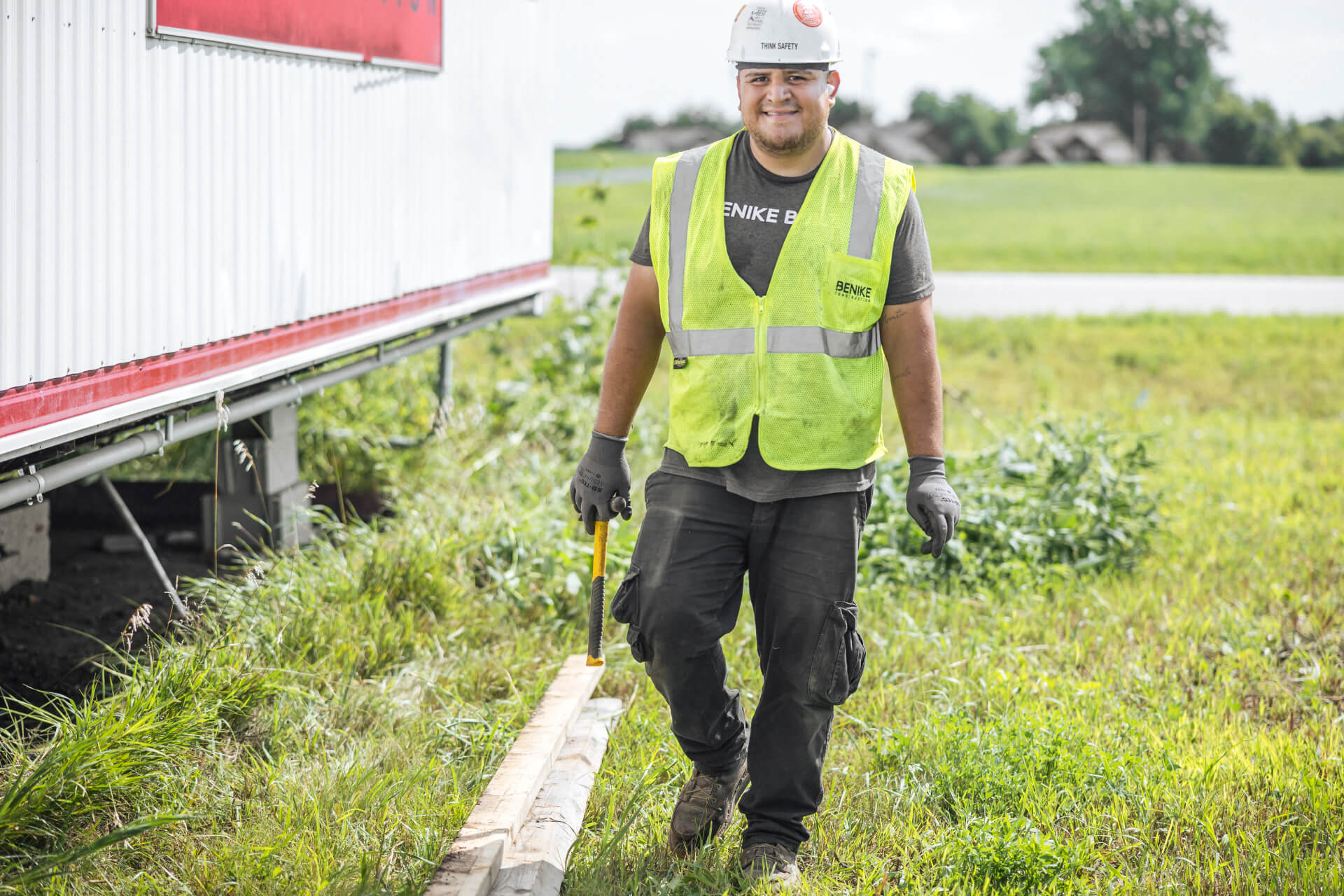 Construction worker in a safety vest and helmet carries a wooden beam outside on grassy ground near a building.