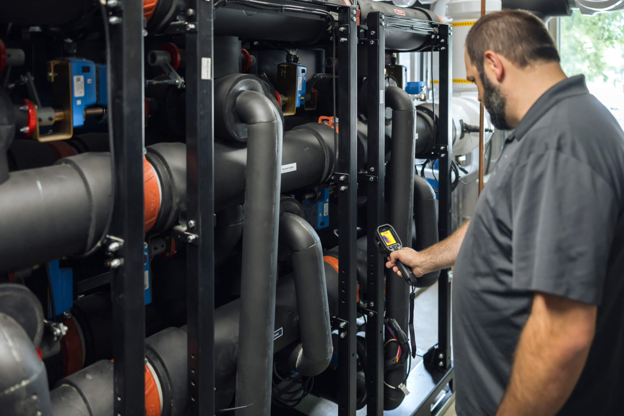 A man uses a handheld device to inspect large black pipes in an industrial setting.