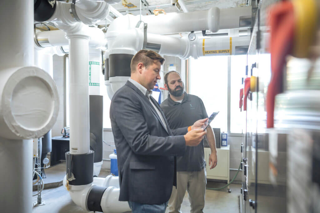 Two men stand in an industrial room with pipes, discussing equipment and reviewing data on a tablet, highlighting their role in managing the built environment.