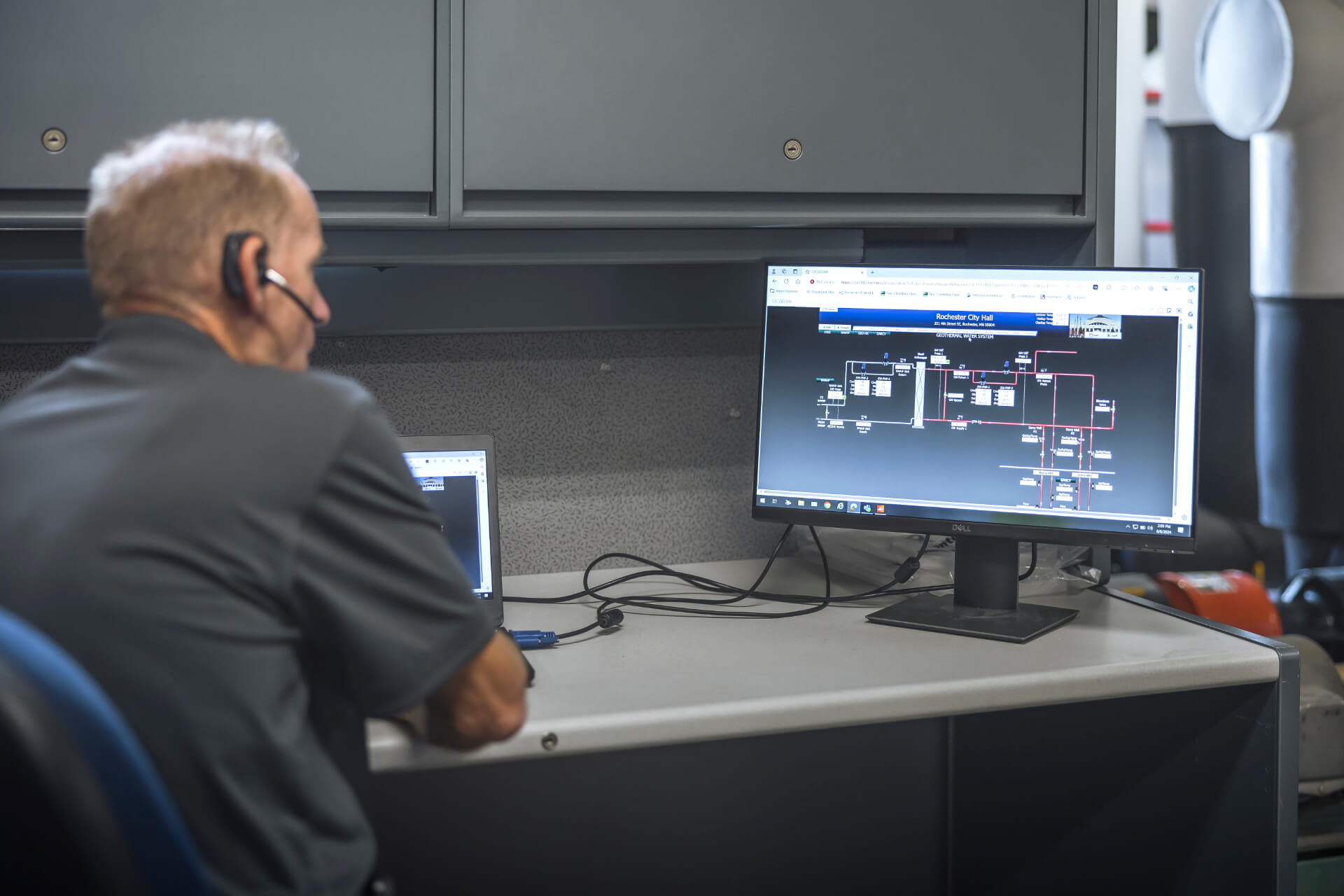 Man with headset sits at desk, looking at a computer screen displaying a technical diagram or system map.