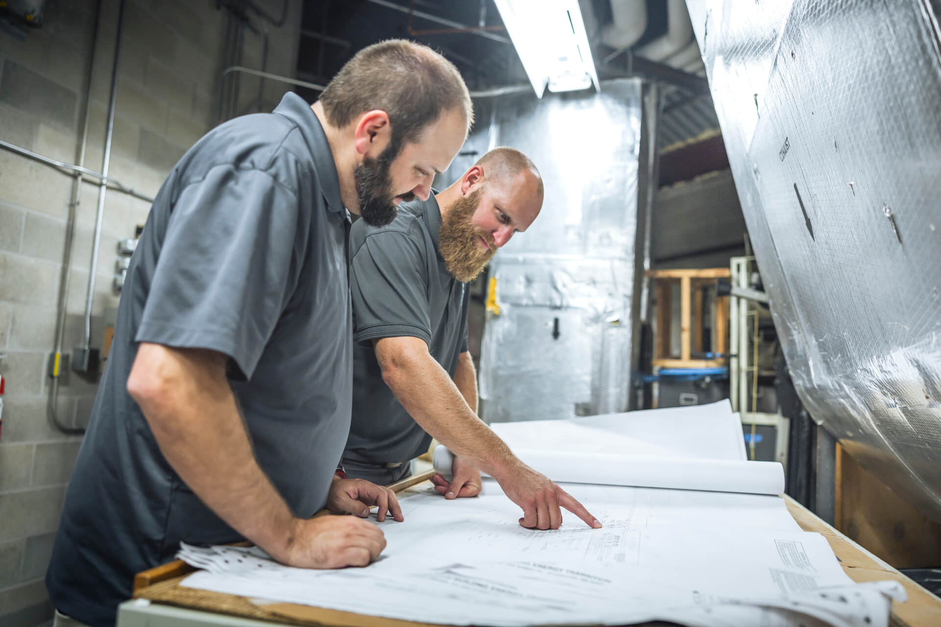 Two men in gray shirts examine and discuss blueprints on a table in an industrial setting.