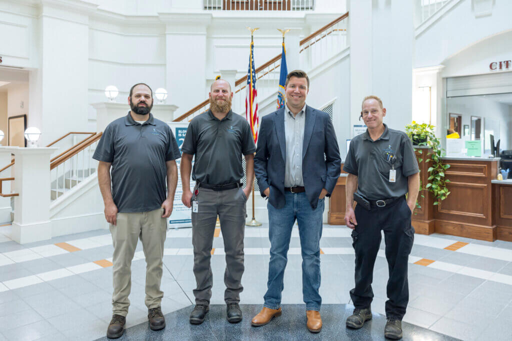 Four men stand indoors in front of American flags, three in work uniforms and one in a blazer and jeans, representing diverse roles within the built environment.