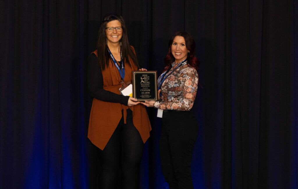 Two women stand smiling, holding a Best Places to Work plaque award together in front of a dark curtain backdrop, celebrating their recognition in Southeast Minnesota.