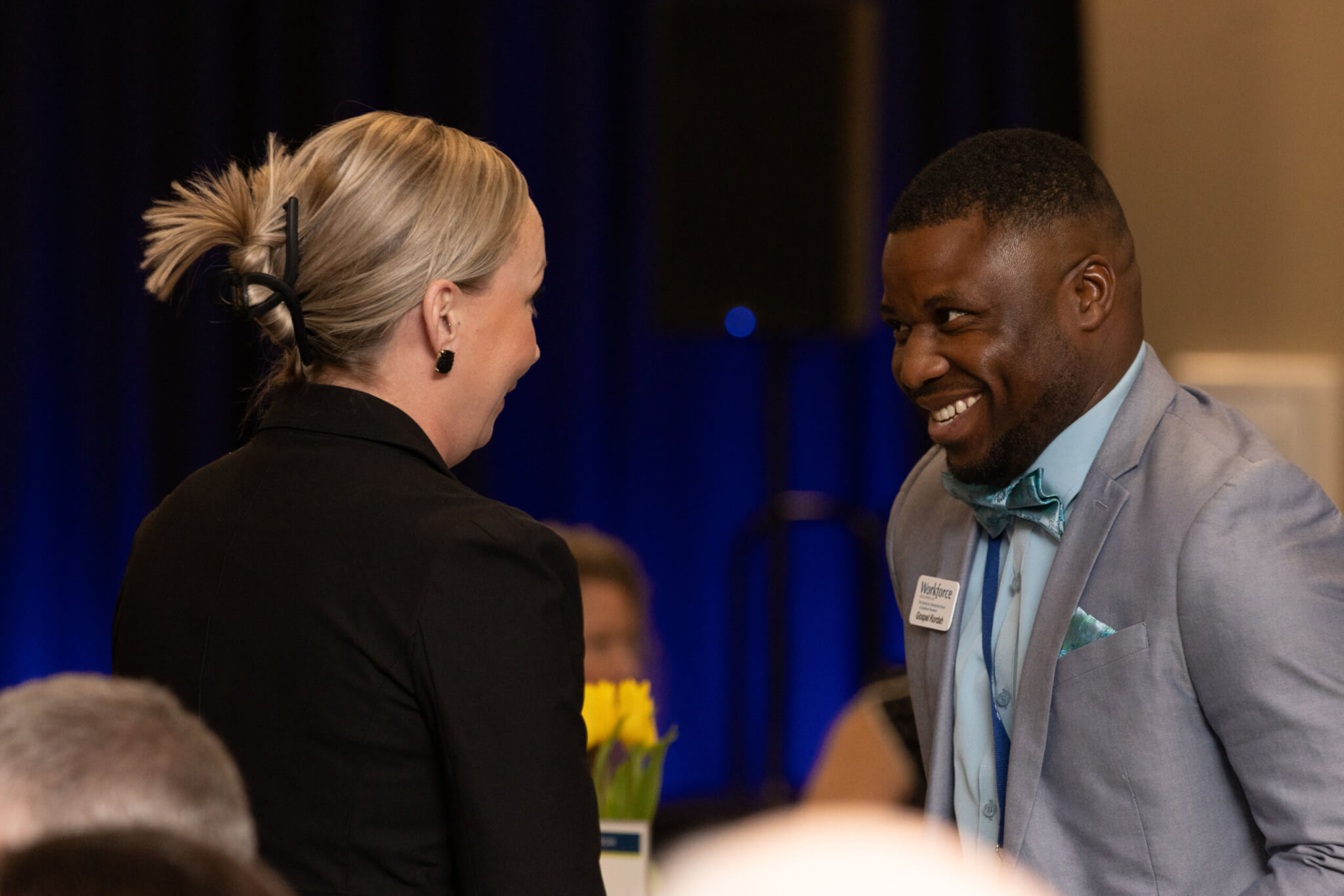 A man in a light suit and bow tie smiles at a woman with blonde hair during an indoor event.