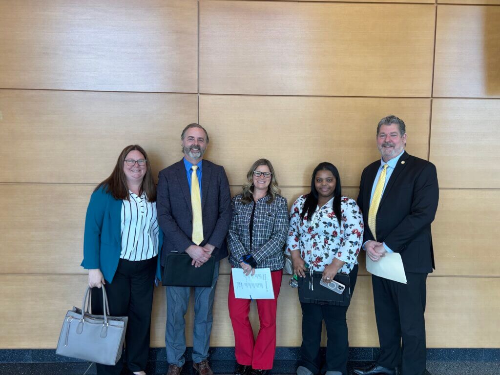 Five young adults stand smiling in business attire against a wooden wall, holding papers and bags.