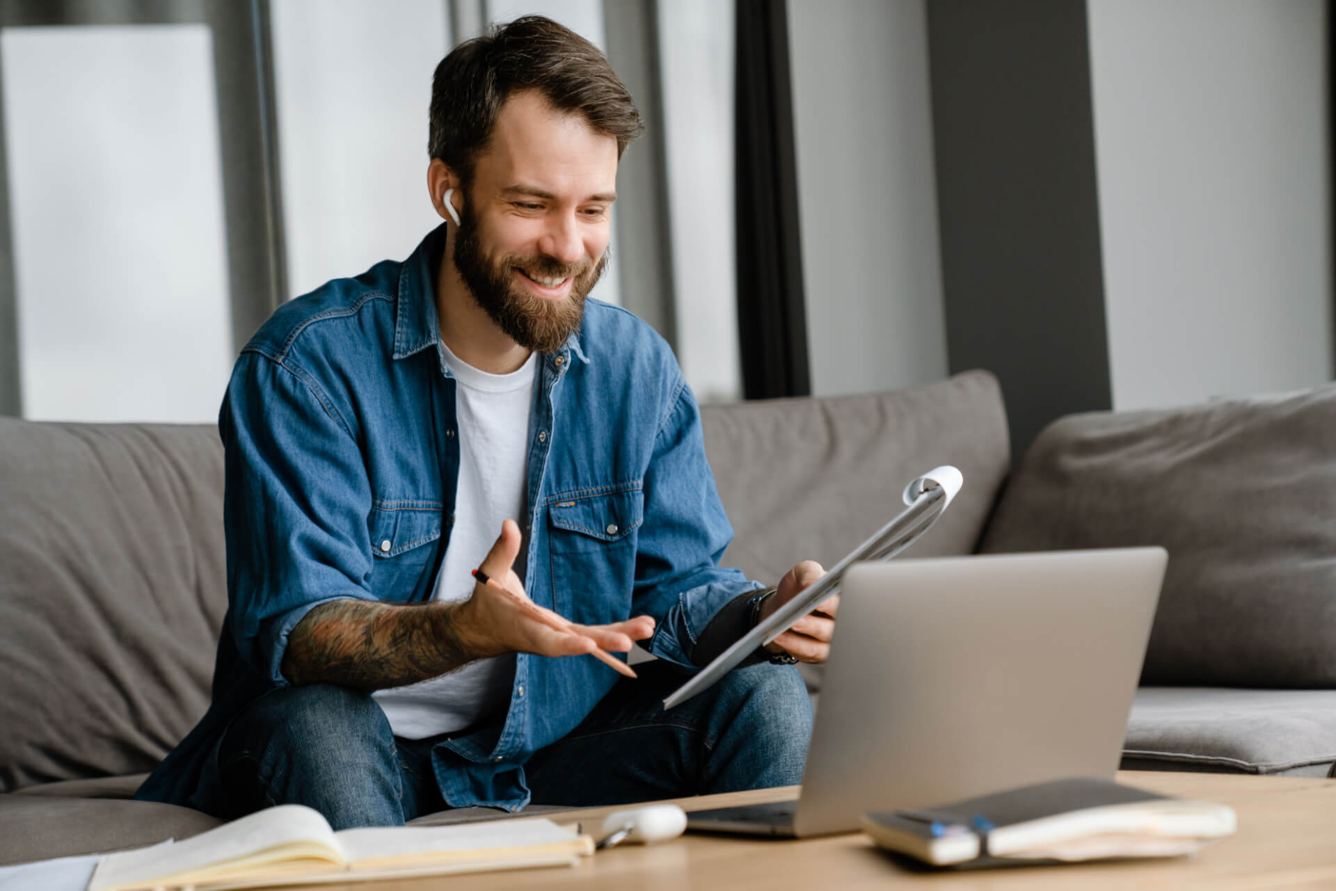 Smiling man on a sofa gestures during a video call on a laptop, holding a clipboard and wearing earbuds.