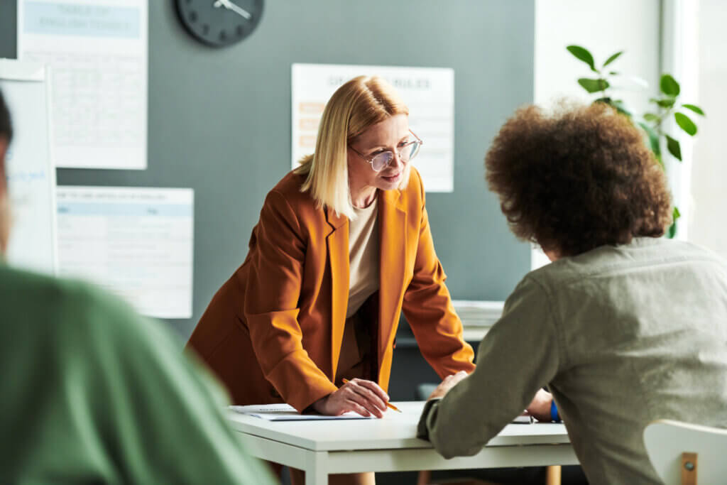 Woman in orange blazer leans over table, talking to seated colleague in a modern office setting.