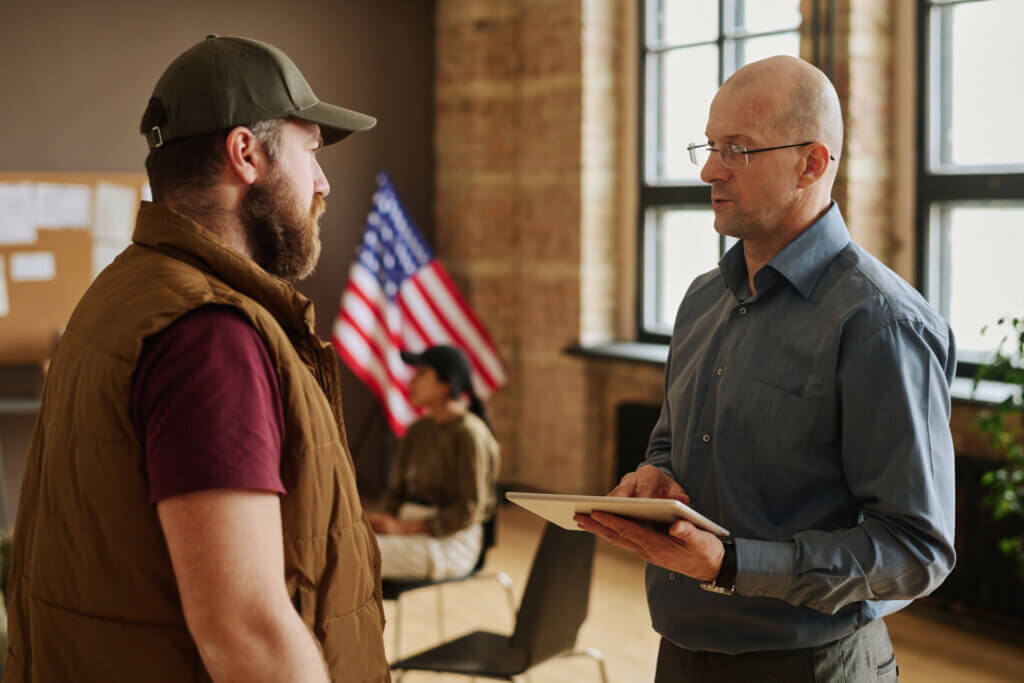 Two men talking indoors, one holding a tablet, with an American flag and a seated person in the background—setting the scene for a discussion about Veteran Services.
