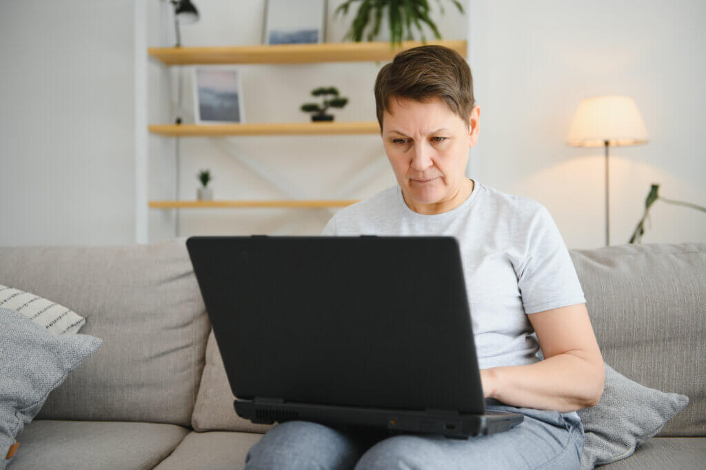 An individual sits on a couch, focused on a laptop in a cozy living room with shelves and plants, possibly exploring Adult Services in the comfort of their home.