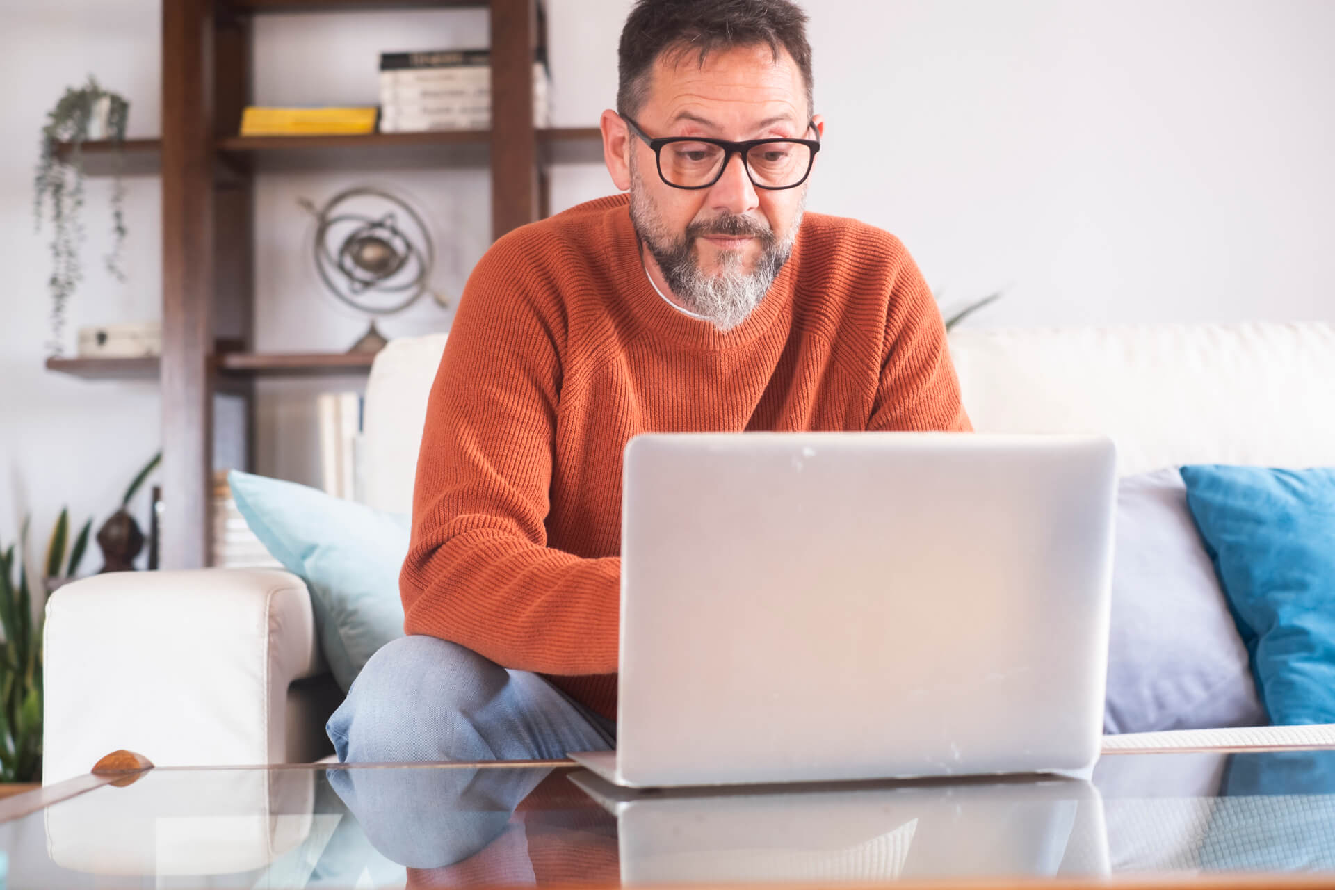 Middle-aged man with glasses using a laptop on a glass table, sitting on a sofa in a cozy living room, browsing Adult Services online.