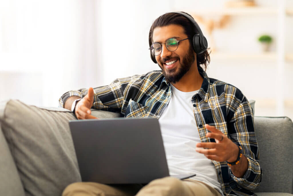 Smiling man with headphones talking during a video call on his laptop while sitting on a couch.