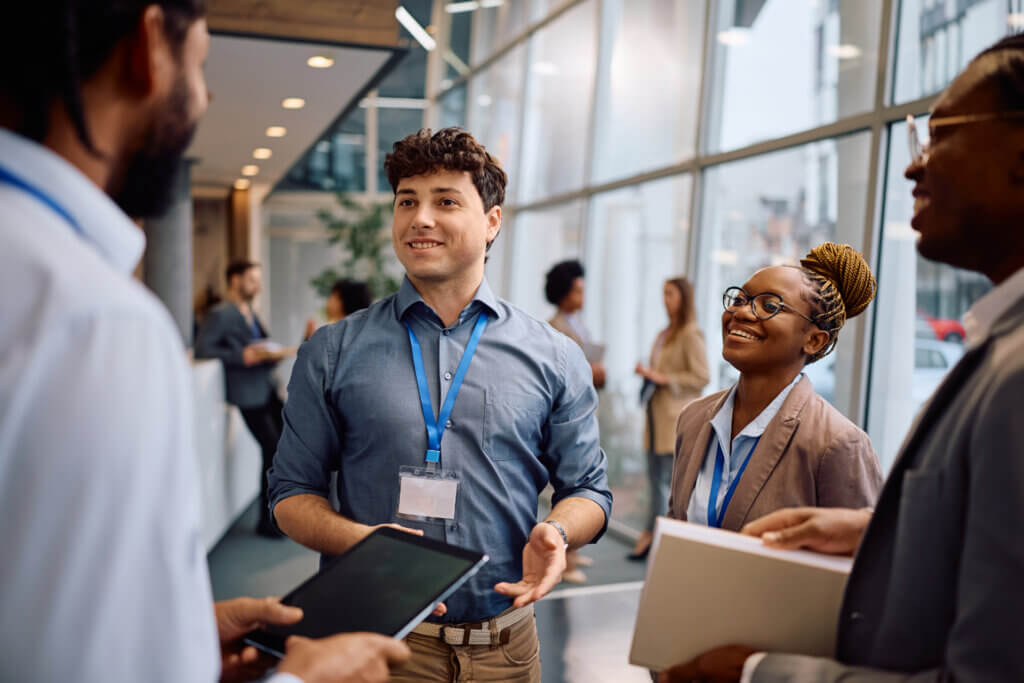Group of professionals wearing name badges chatting and smiling in a modern office hallway, showcasing a positive environment focused on recruitment and retention assistance.