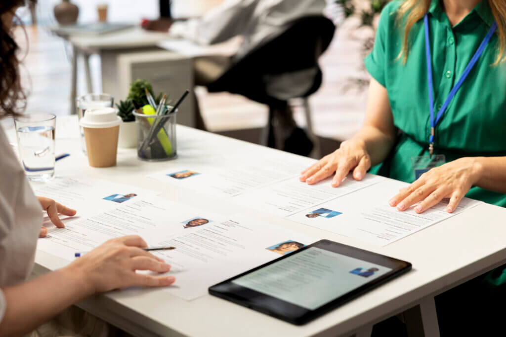 Two people review resumes at a desk with documents, a tablet, and office supplies.