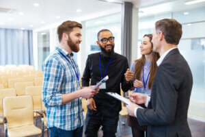 Four people with name badges talking and smiling in an empty conference room with chairs and glass walls during staff training.