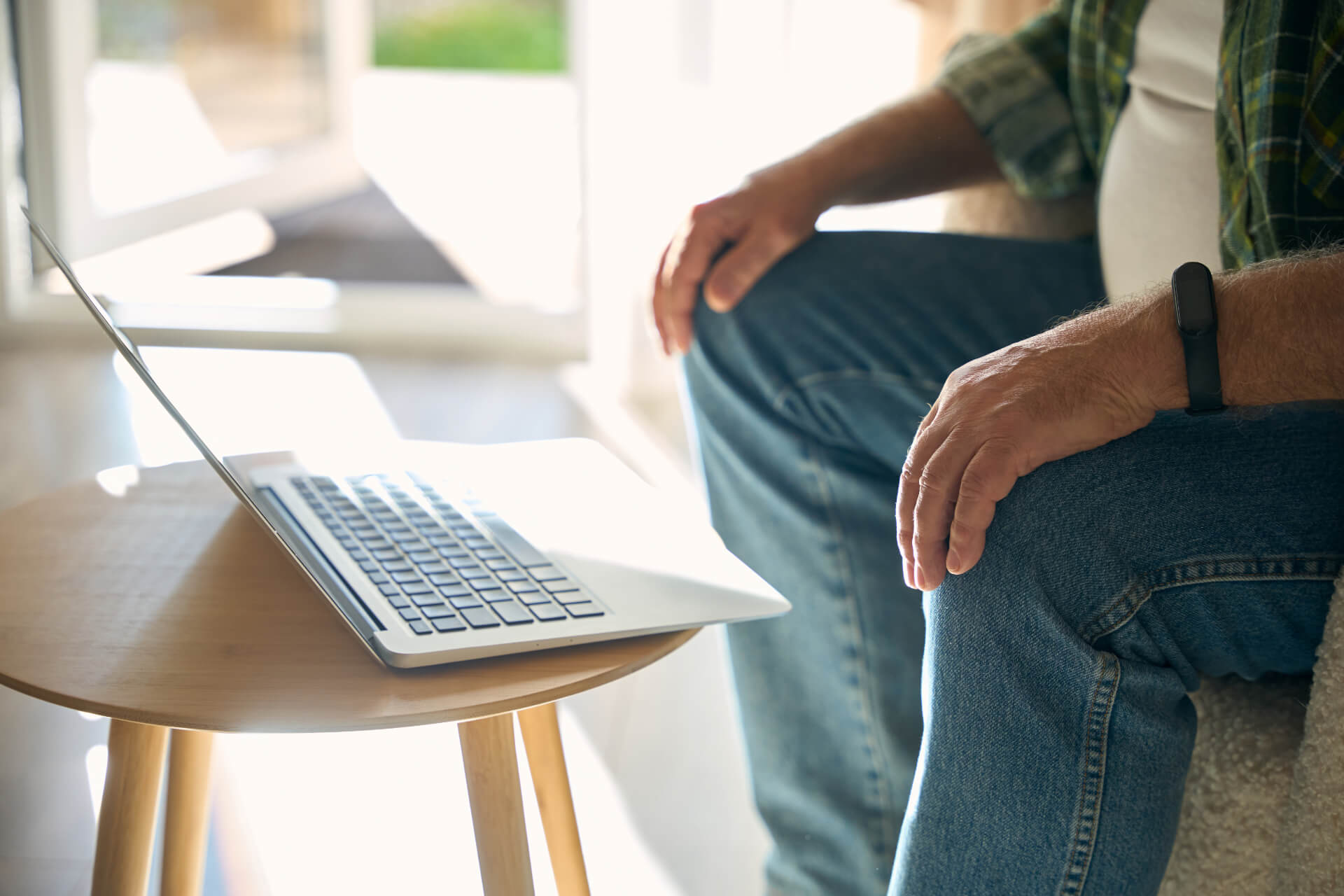 A person in jeans sits beside a laptop on a small table, sunlight streaming through a nearby window, suggesting a calm moment before engaging with adult services online.