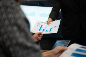 Close-up of hands exchanging a Labor Market Information bar chart printout in a business meeting, with graphs displayed on a screen in the background.