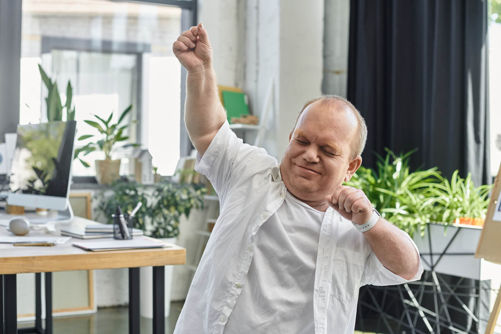A man in a white shirt dances joyfully in a bright office with lots of green plants.