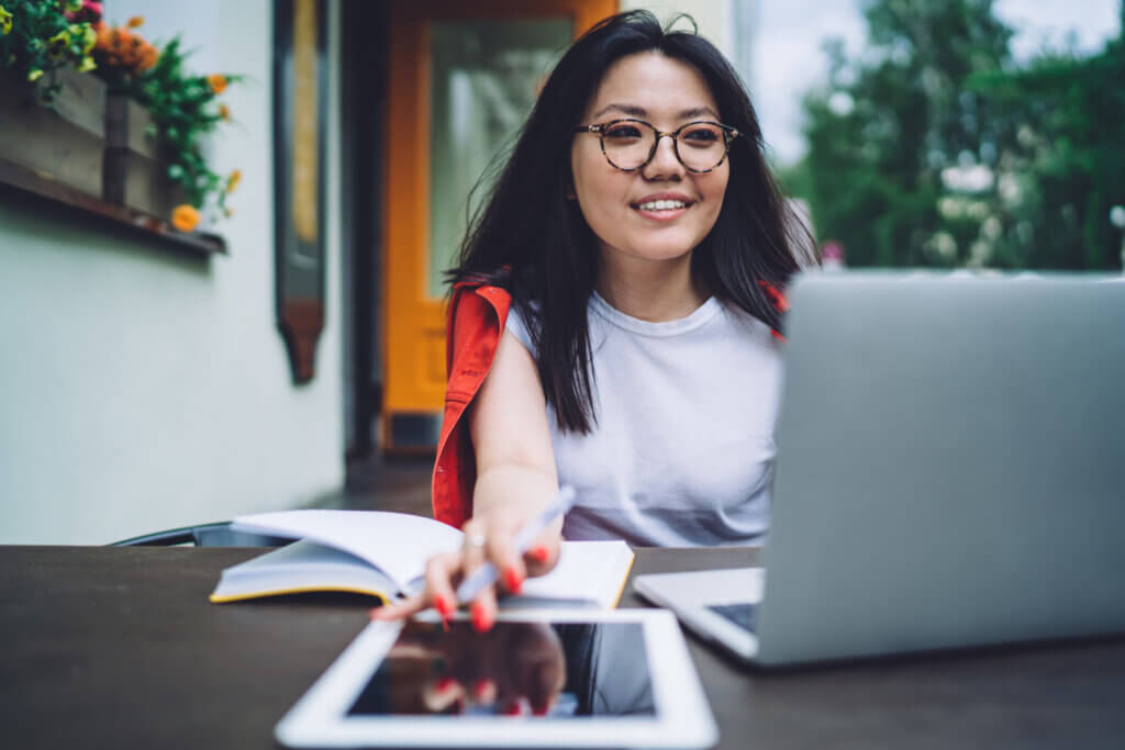 Woman smiling while working on a laptop outdoors, with an open notebook and tablet on the table.