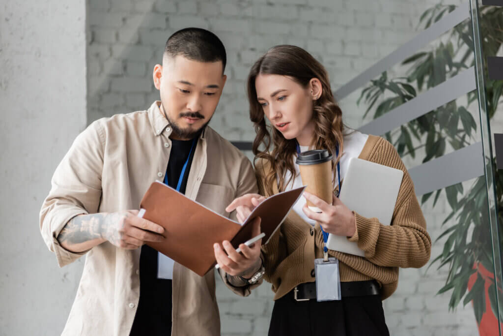 Two colleagues reviewing documents together; one holds a folder, the other a laptop and coffee cup.