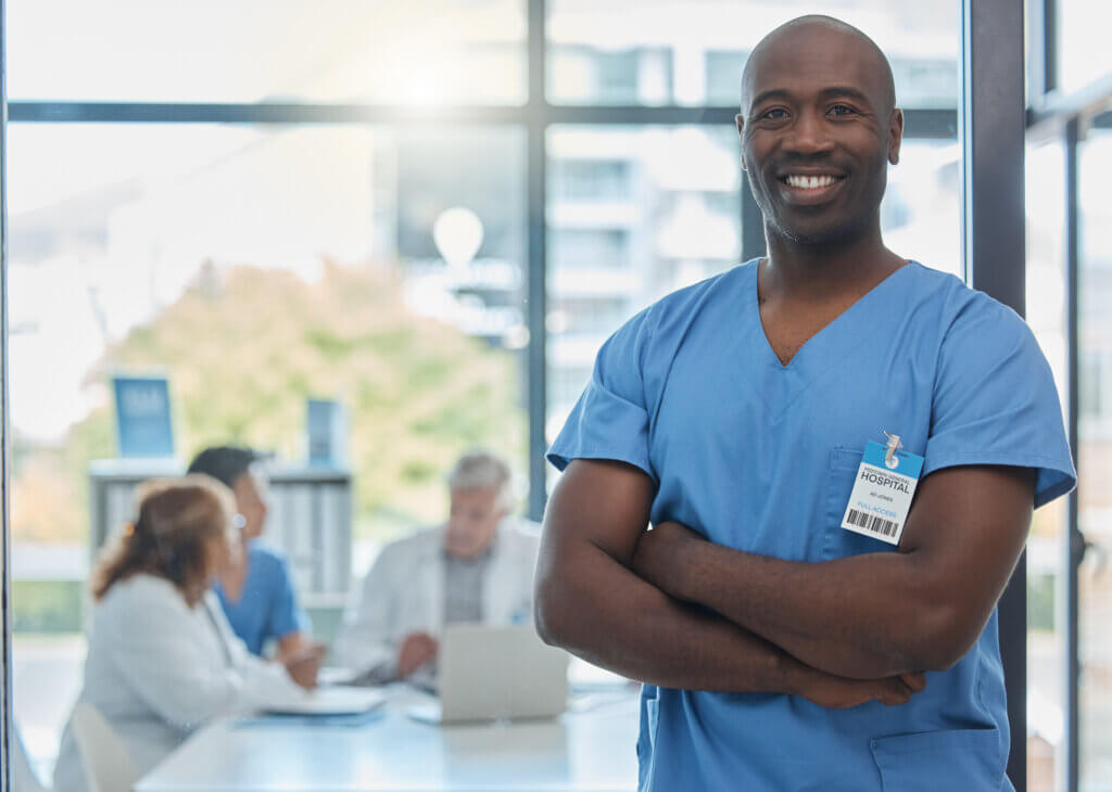 Smiling healthcare worker in blue scrubs stands with arms crossed, showcasing the rewarding career pathways available in healthcare, while colleagues meet in the background.