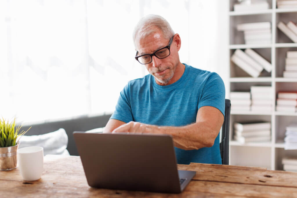Older man in glasses and blue shirt works on a laptop at a wooden table in a bright room, researching Adult Services.