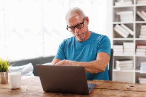 Older man in glasses and blue shirt works on a laptop at a wooden table in a bright room, researching Adult Services.