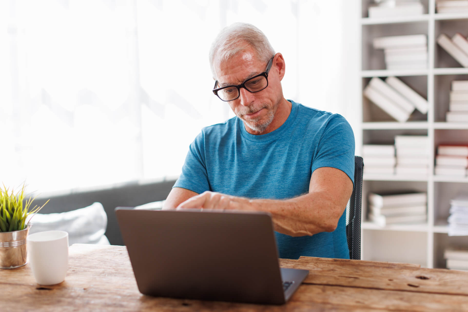 Older man in glasses and blue shirt works on a laptop at a wooden table in a bright room, researching Adult Services.